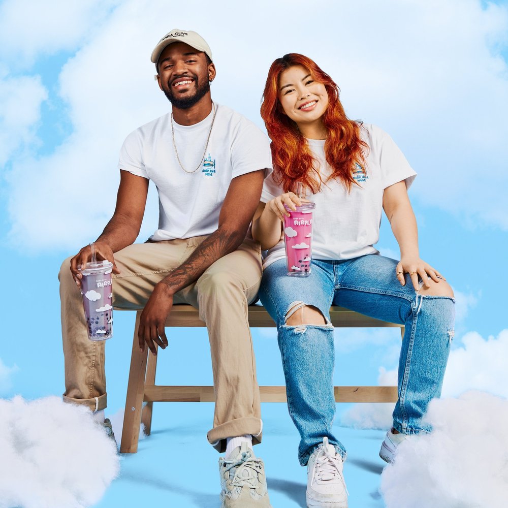 Man and woman sitting on a bench smiling wearing airplane mode t-shirts and holding drink tumblers